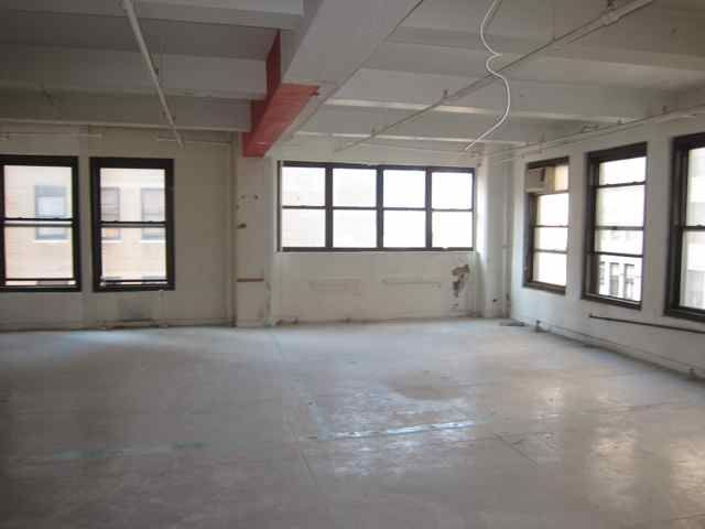 Empty, well-lit commercial loft rental with large windows. Bare floor, exposed pipes, worn white walls with peeling paint.