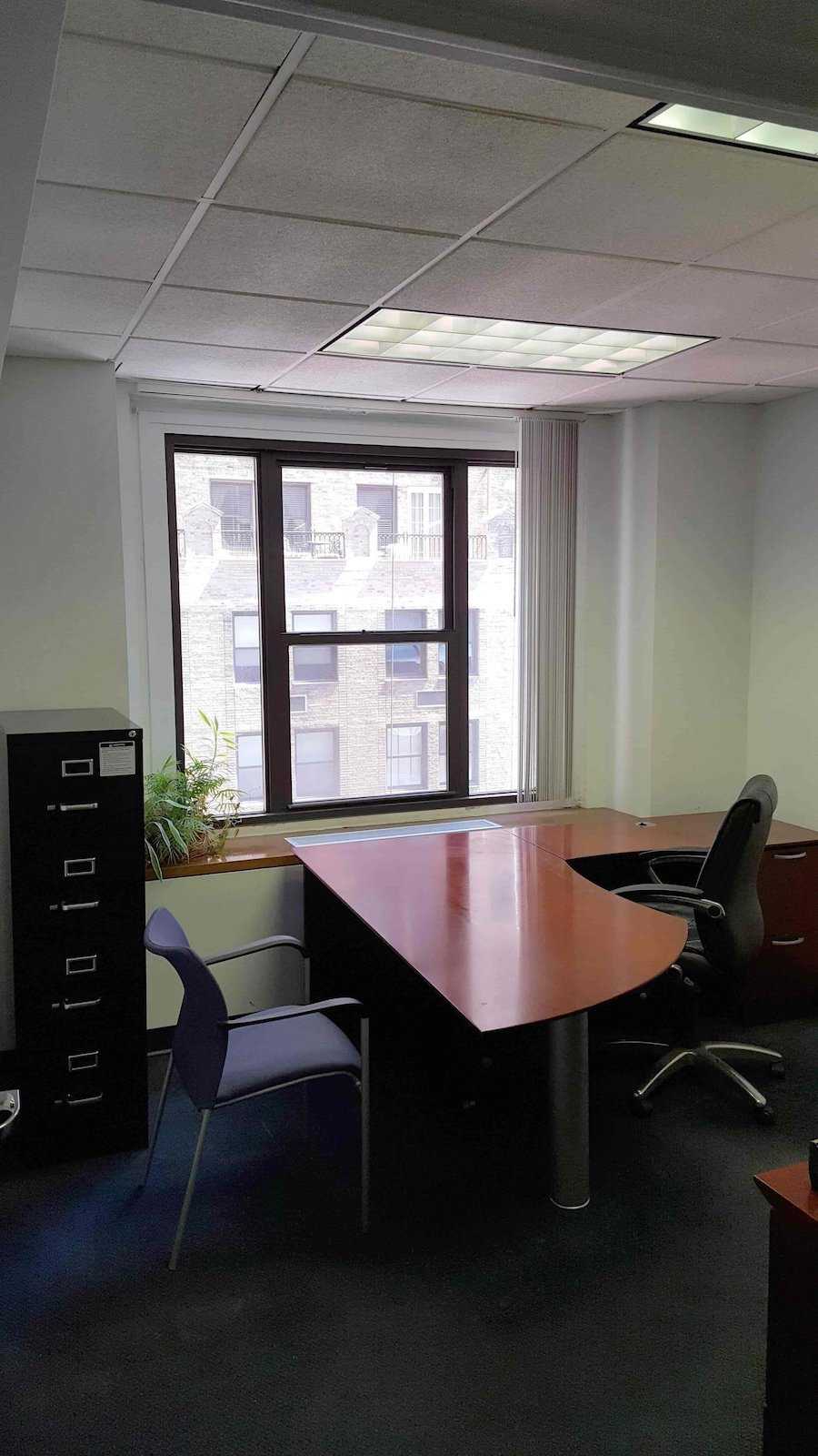 A well-lit office with wooden desk, black chair, purple guest chair, filing cabinet, and potted plant.