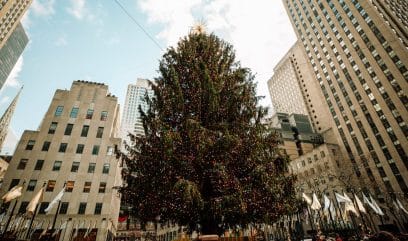 The iconic Rockefeller Christmas Tree against NYC skyscrapers on a sunny winter day.